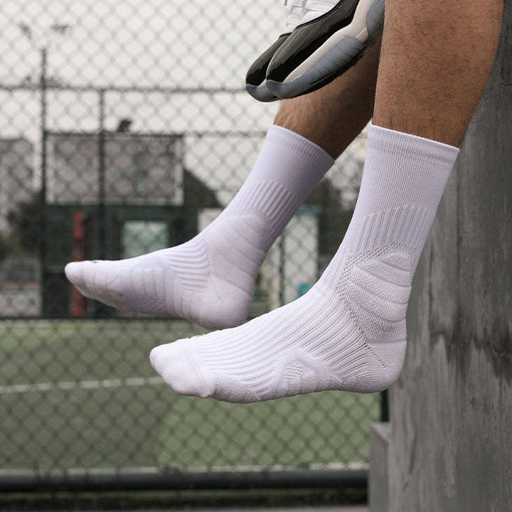 An athlete wearing white crew socks sitting on a ledge with a tennis racket in hand against a chain-link fence backdrop.